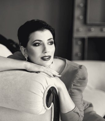 Black-and-white portrait of a glamorous dark-haired woman with bold lips, leaning on a vintage sofa