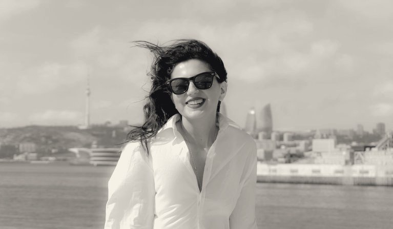 Black-and-white photo of a smiling woman in a crisp white shirt and dark sunglasses, hair blowing in the wind by the sea