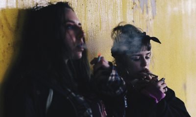 two women smoking while leaning on yellow wall